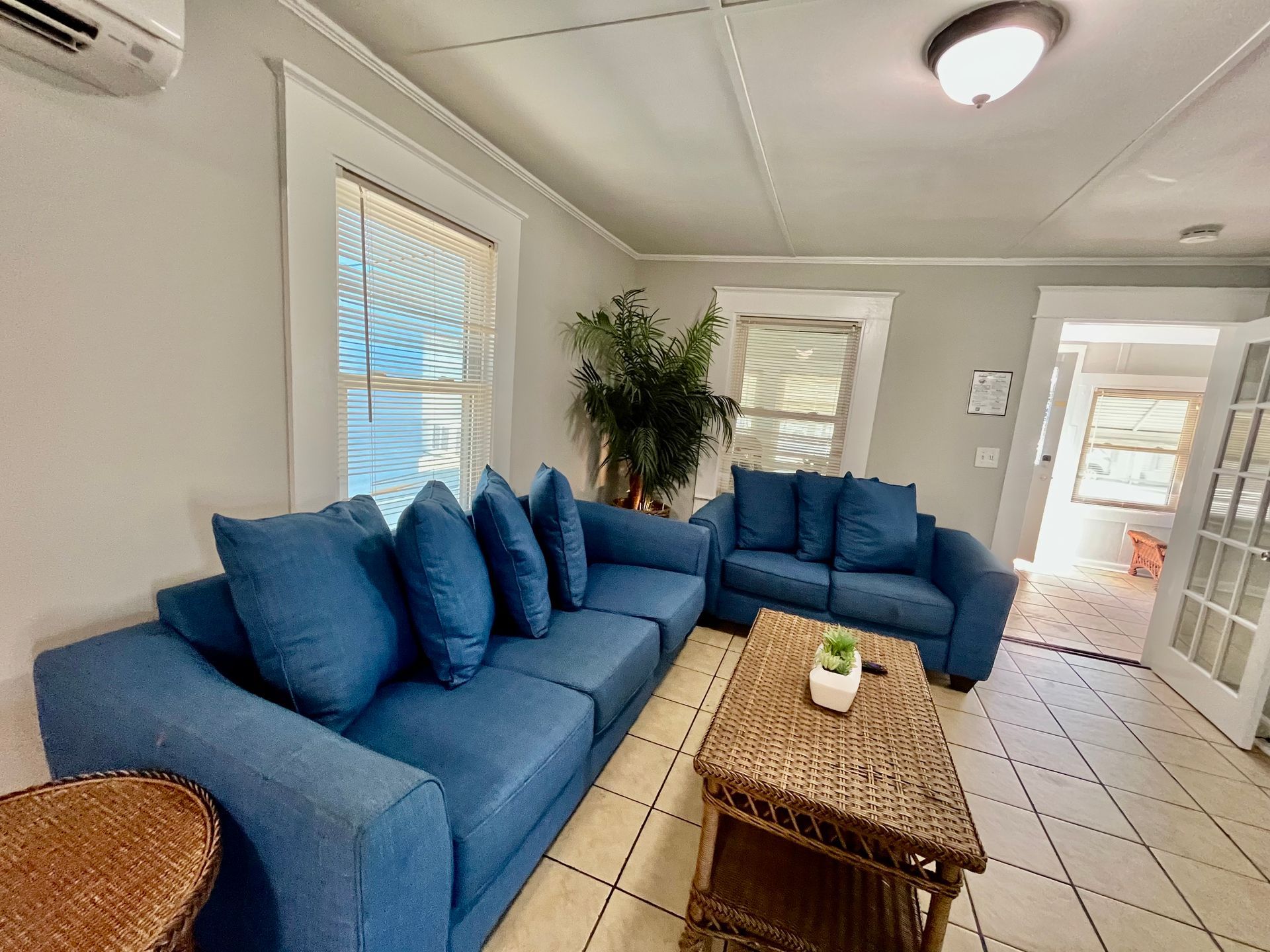 A spacious living area at Seabirds Motel at Kure Beach with a large blue sectional sofa, wicker coffee table, and decorative plants. This room provides a comfortable gathering space for families and groups, enhancing the pet-friendly lodging experience in North Carolina.