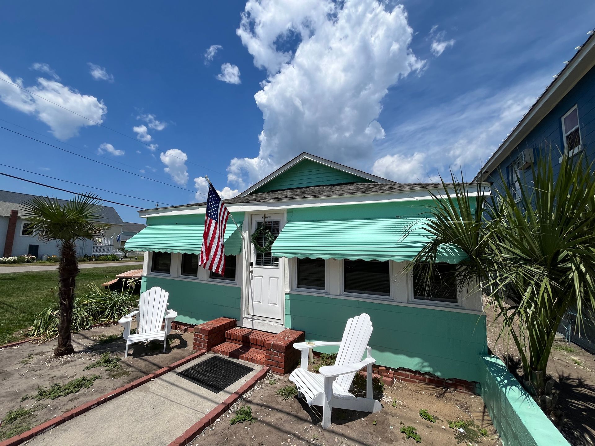 The exterior of Seabirds Motel at Kure Beach showcasing a charming teal-painted building with white trim, a U.S. flag, and outdoor seating under sunny blue skies. This inviting setting reflects the motel's welcoming and pet-friendly atmosphere in North Carolina.