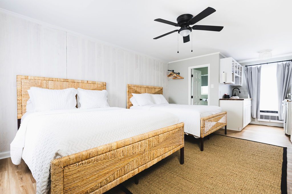 A bright and airy guest room at Seabirds Motel at Kure Beach featuring two comfortable beds with white linens, a woven rattan bed frame, a ceiling fan, and light-colored furnishings. The room's spacious layout and additional bed make it perfect for families or groups seeking a pet-friendly lodging option in North Carolina.