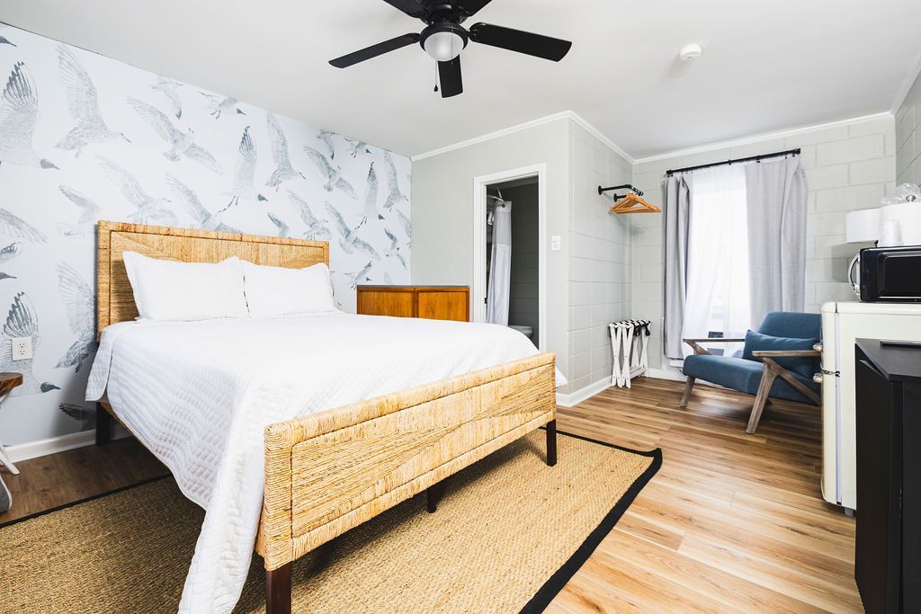 A bright and airy guest room at Seabirds Motel at Kure Beach, featuring a queen-sized bed with a rattan headboard, white linens, and coastal-themed wallpaper. The room's light and inviting atmosphere make it a perfect choice for a relaxing, pet-friendly stay in North Carolina.