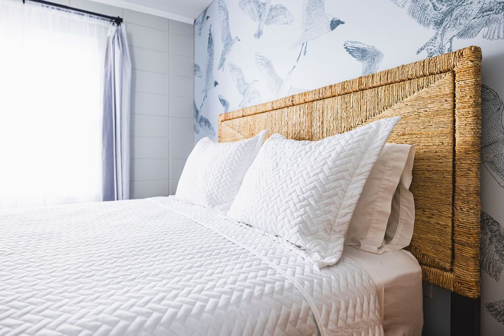 A charming bedroom at Seabirds Motel at Kure Beach featuring a queen-sized bed with a woven rattan headboard, soft white bedding, and subtle floral wallpaper. This cozy room offers a peaceful and pet-friendly lodging option in North Carolina.