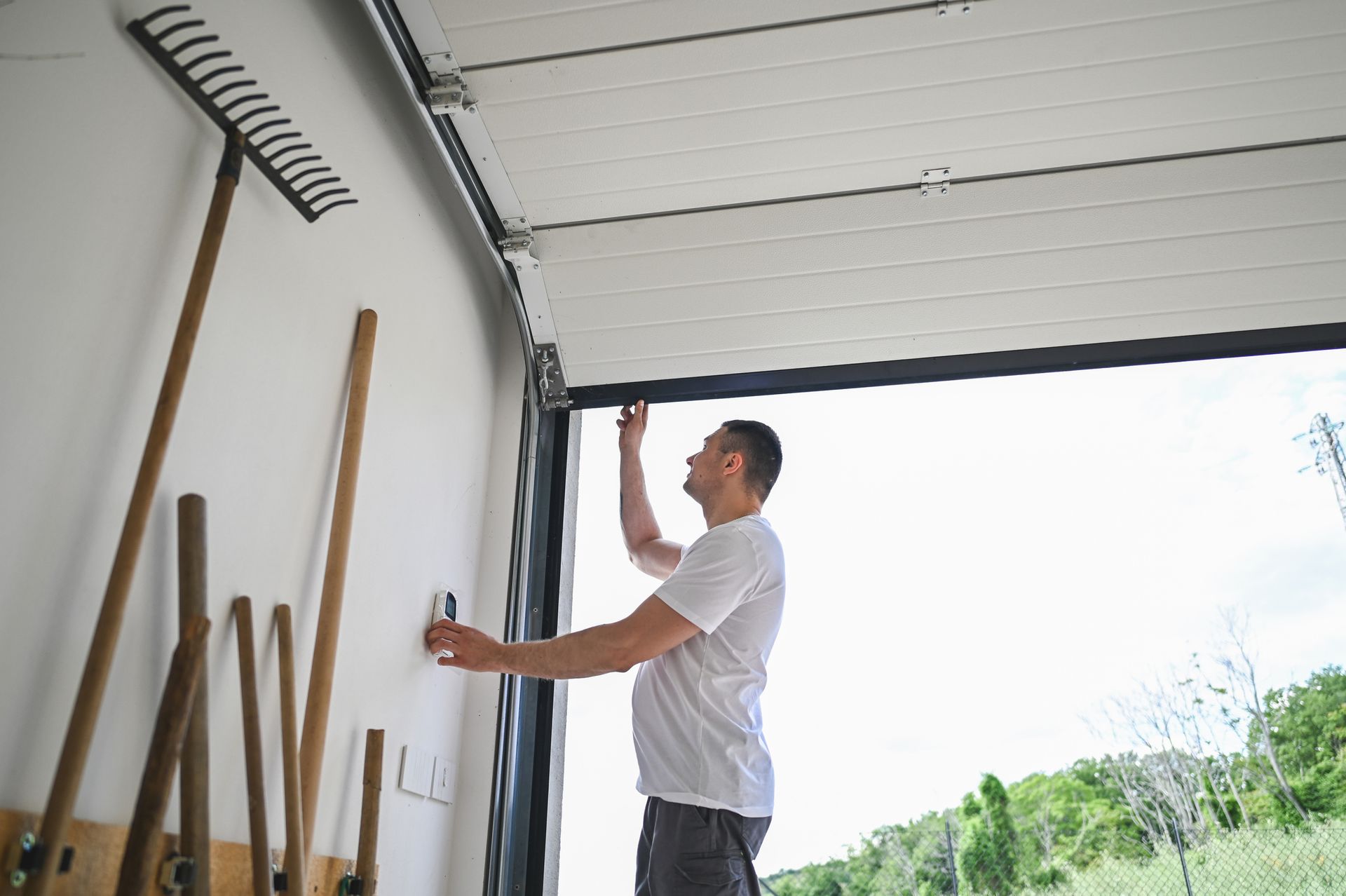 A man is opening a garage door with a rake hanging on the wall.