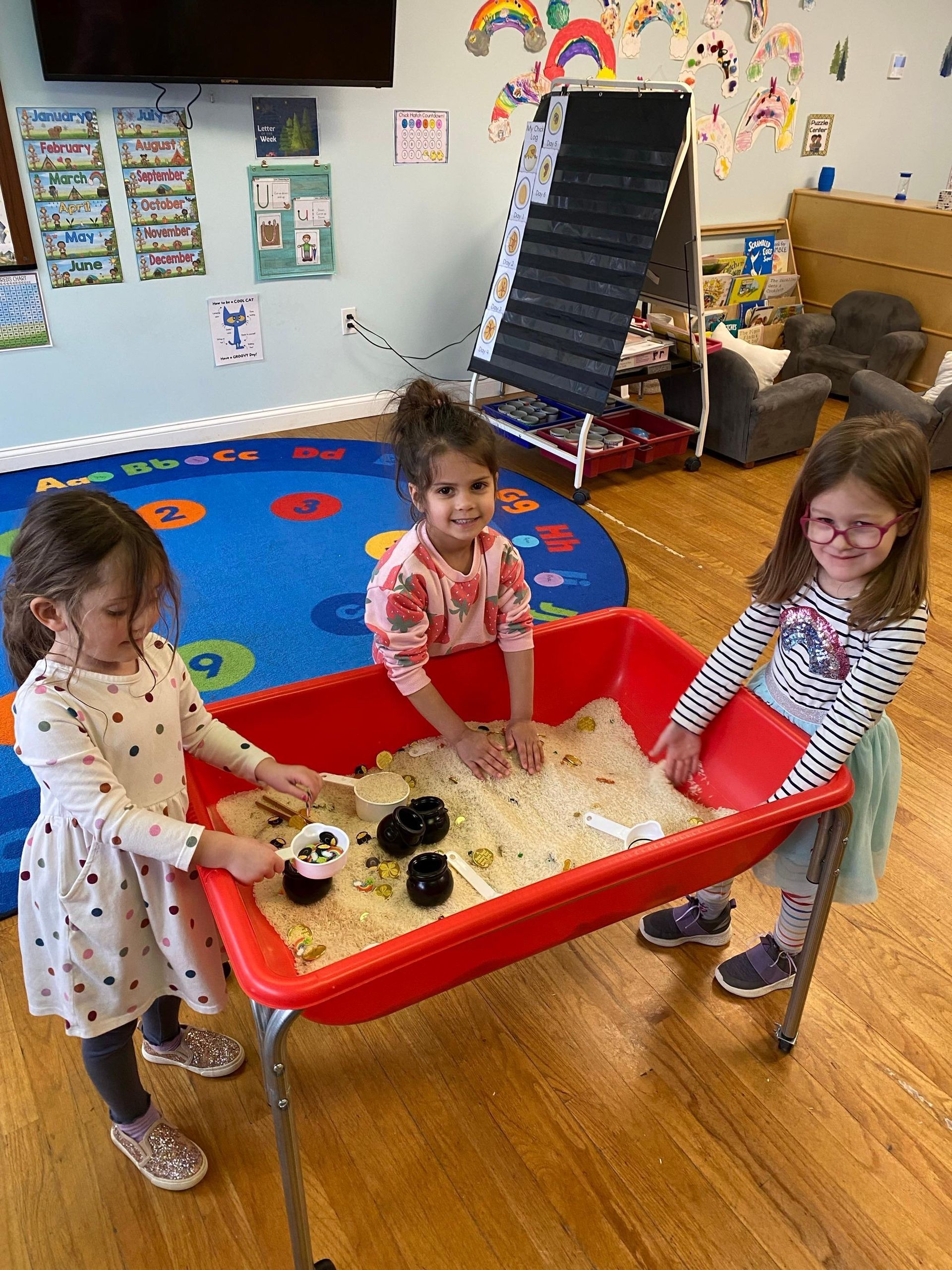 Three young girls are playing in a sandbox in a classroom.