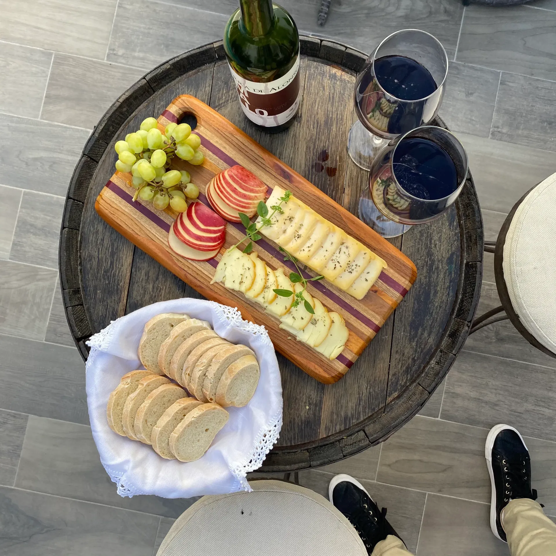 Outdoor snack spread on a round table with grapes, sliced fruit, cheese, wine, and crackers.