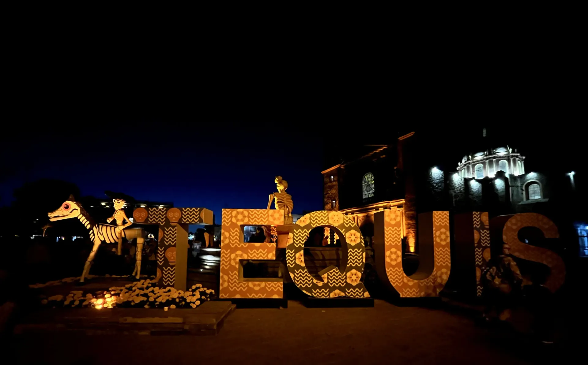 Letrero iluminado de “EQUIS” por la noche con edificios iluminados al fondo.