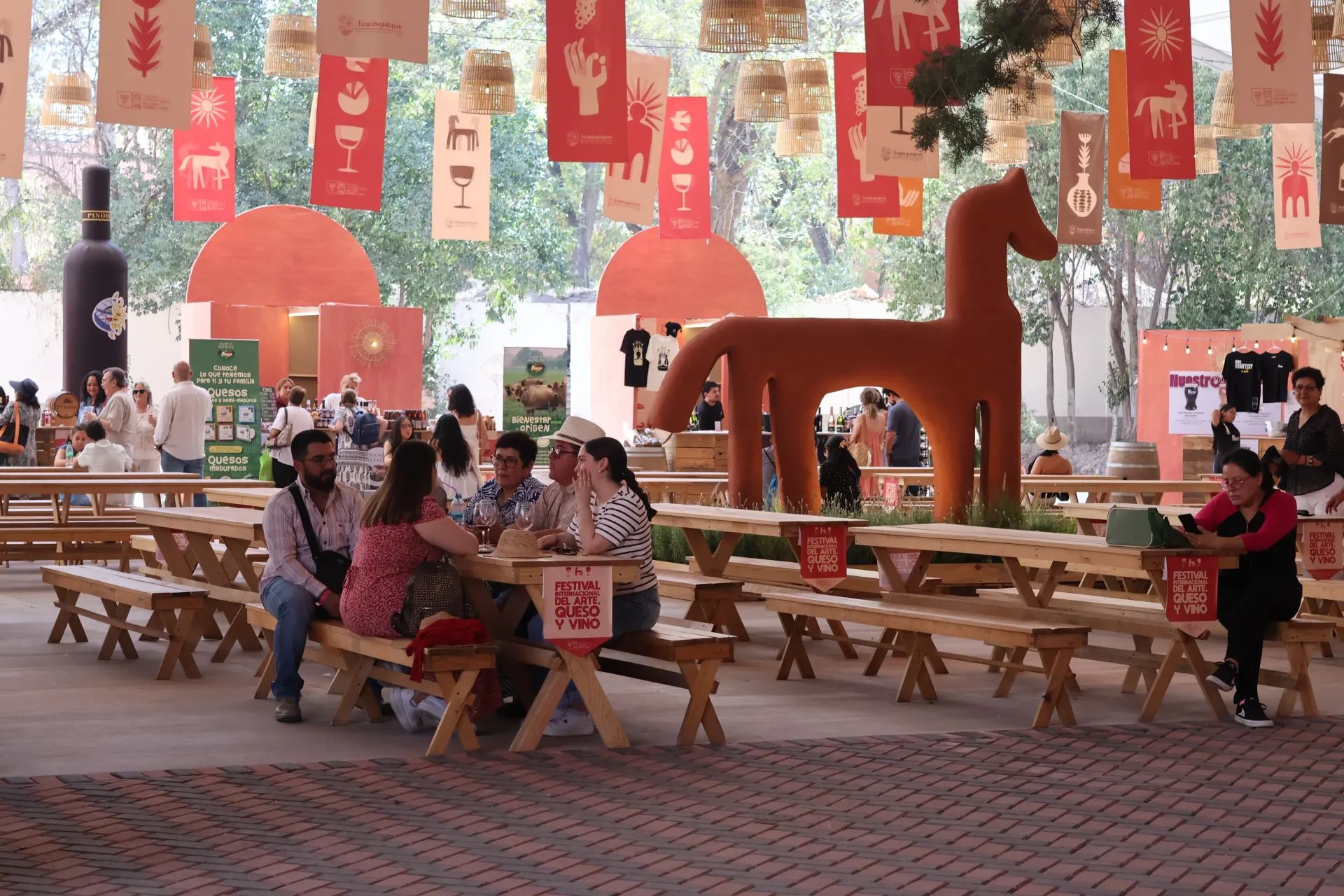 Gente sentada en mesas de picnic en una plaza al aire libre con un ambiente festivo, adornada con pancartas rojas.