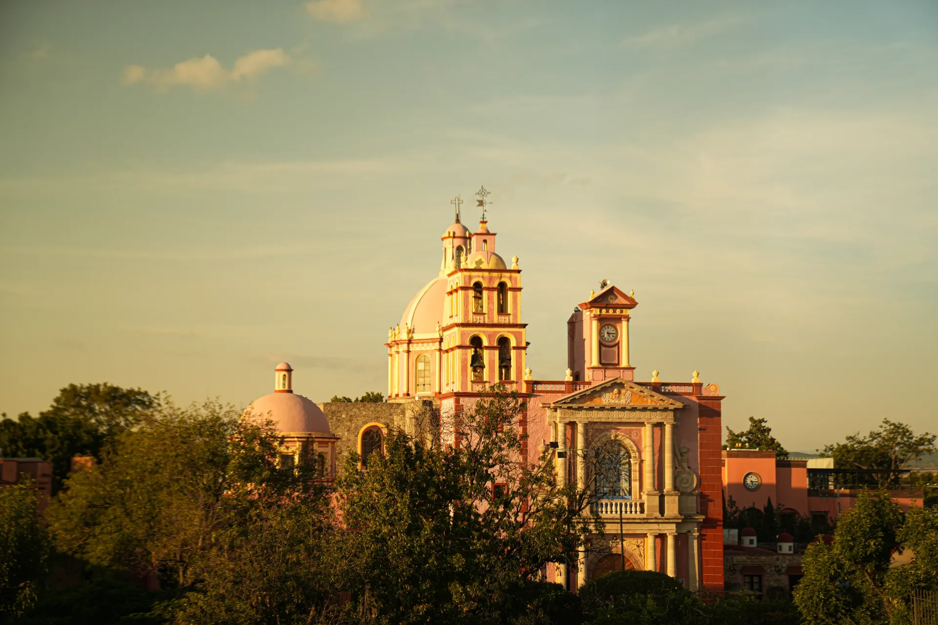 Catedral histórica bañada por el sol, con cúpulas y campanario sobre los árboles al atardecer.