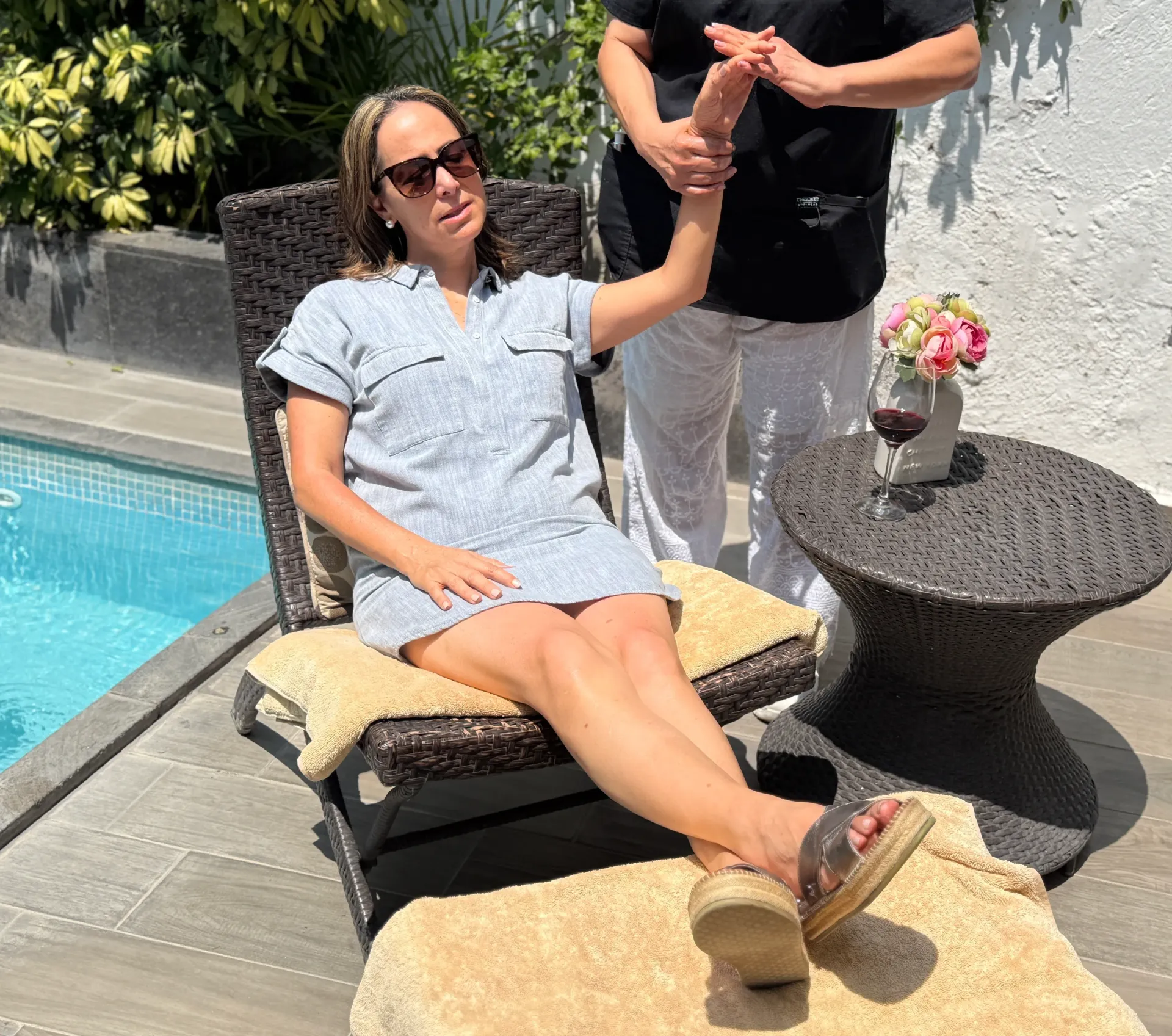 Woman relaxing by a pool in a sun chair, holding hands with someone standing beside her.