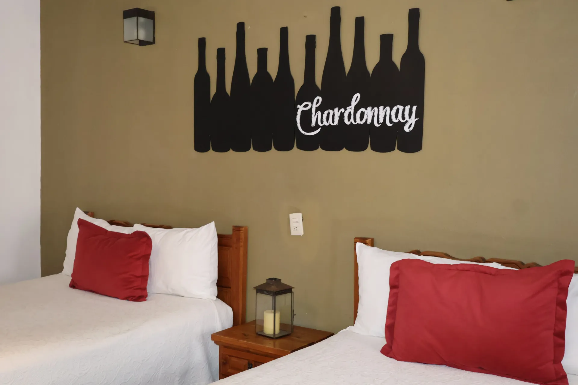 Habitación de hotel con dos camas blancas, almohadas rojas y una botella de Chardonnay como decoración de pared.