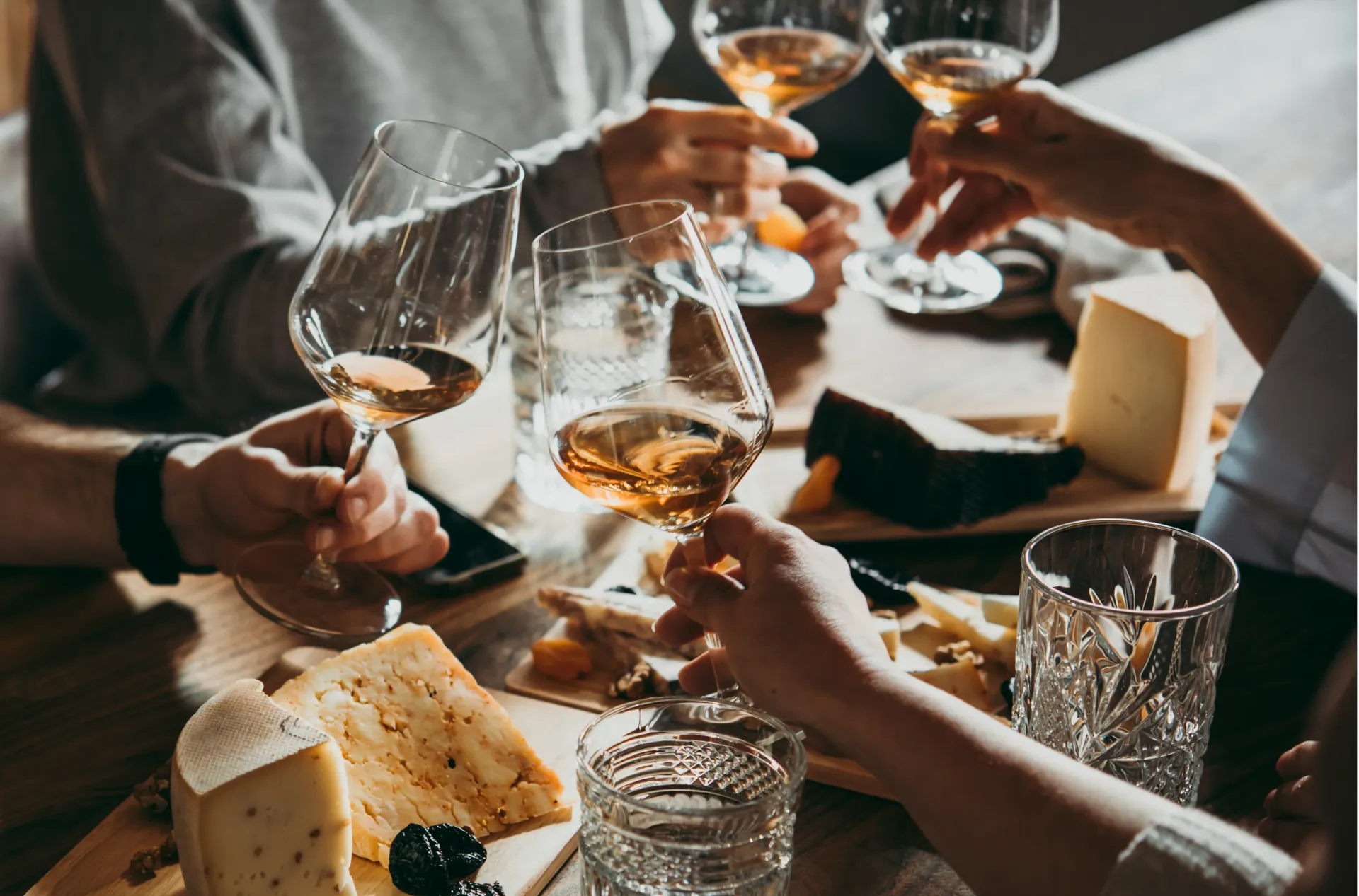 Manos alzando copas de vino sobre una mesa con pan y queso durante una comida compartida.