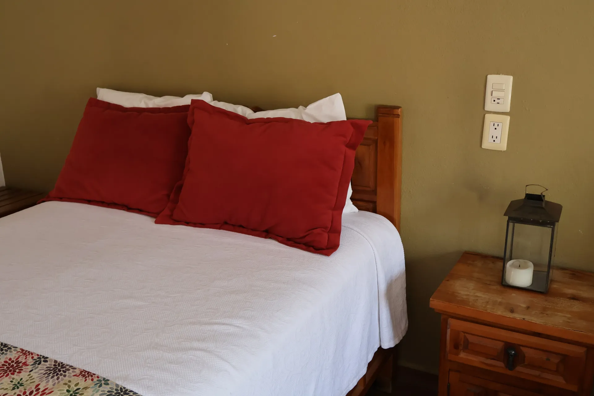 A bed with red pillows and white bedding beside a wooden nightstand with a lantern.