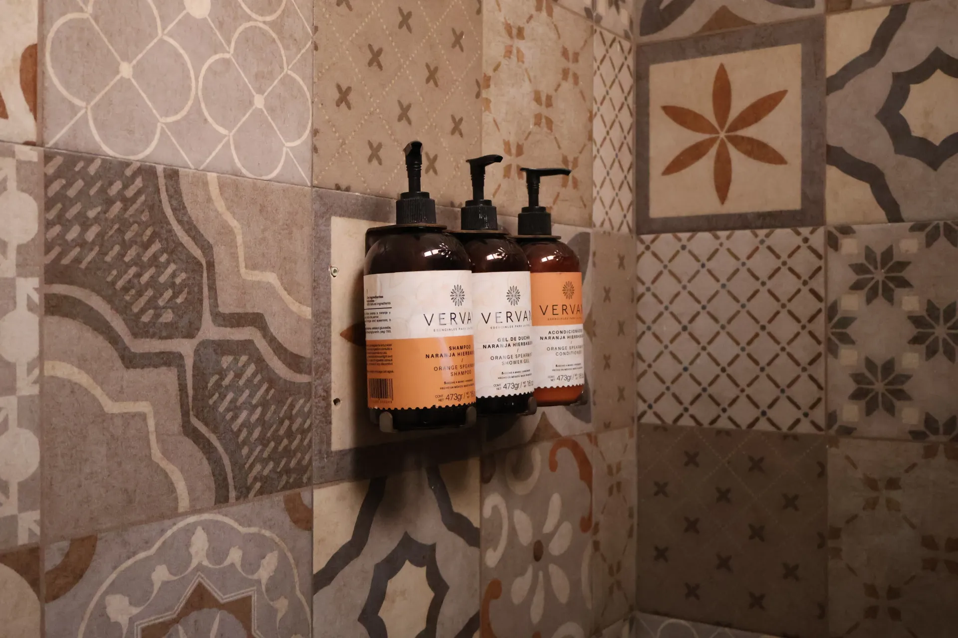 Three pump soap bottles on a wall-mounted shelf in a patterned tiled shower.