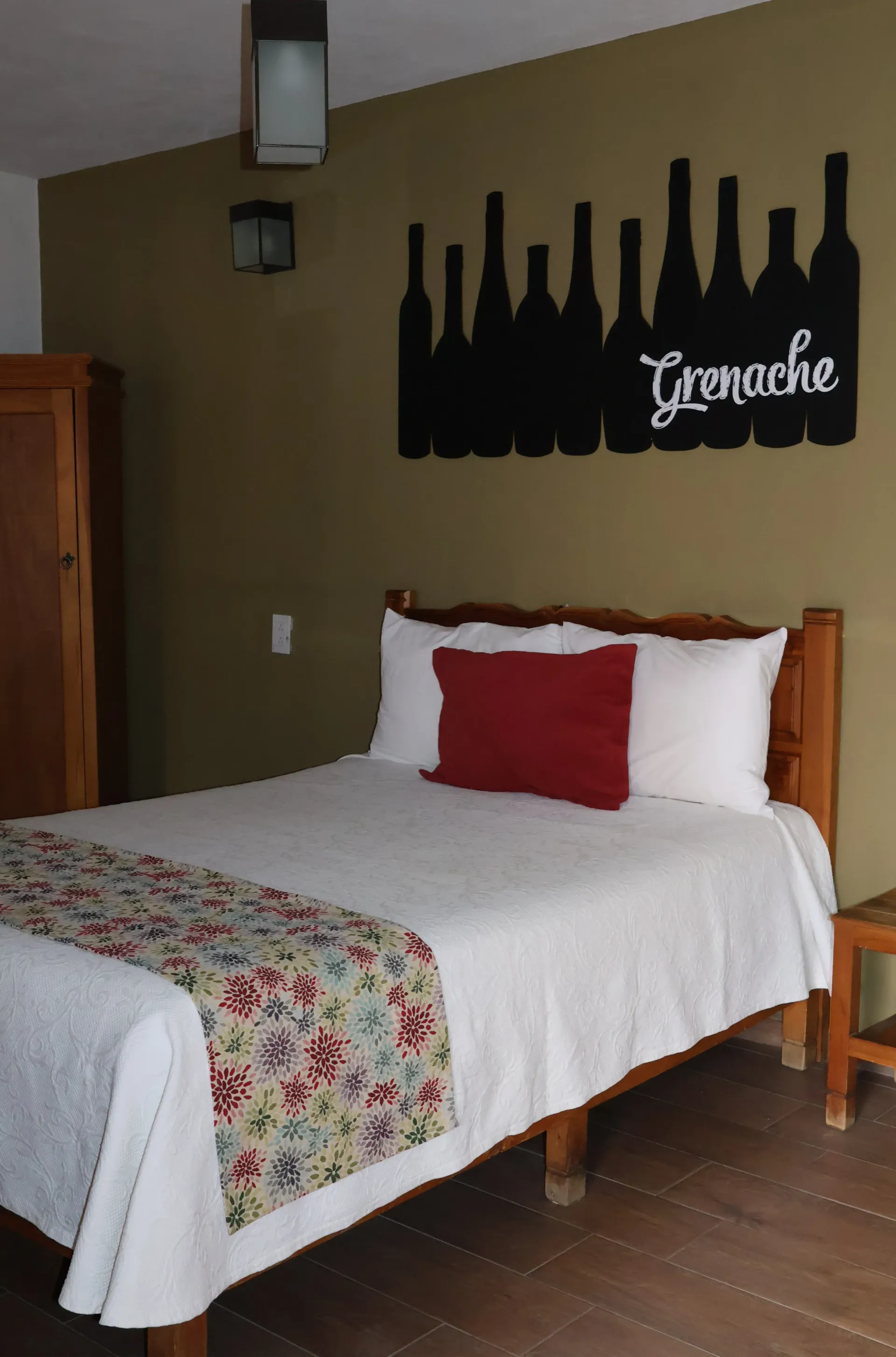 Acogedor dormitorio con cama de madera, camino de cama floral, almohada roja y cuadro con forma de botella.