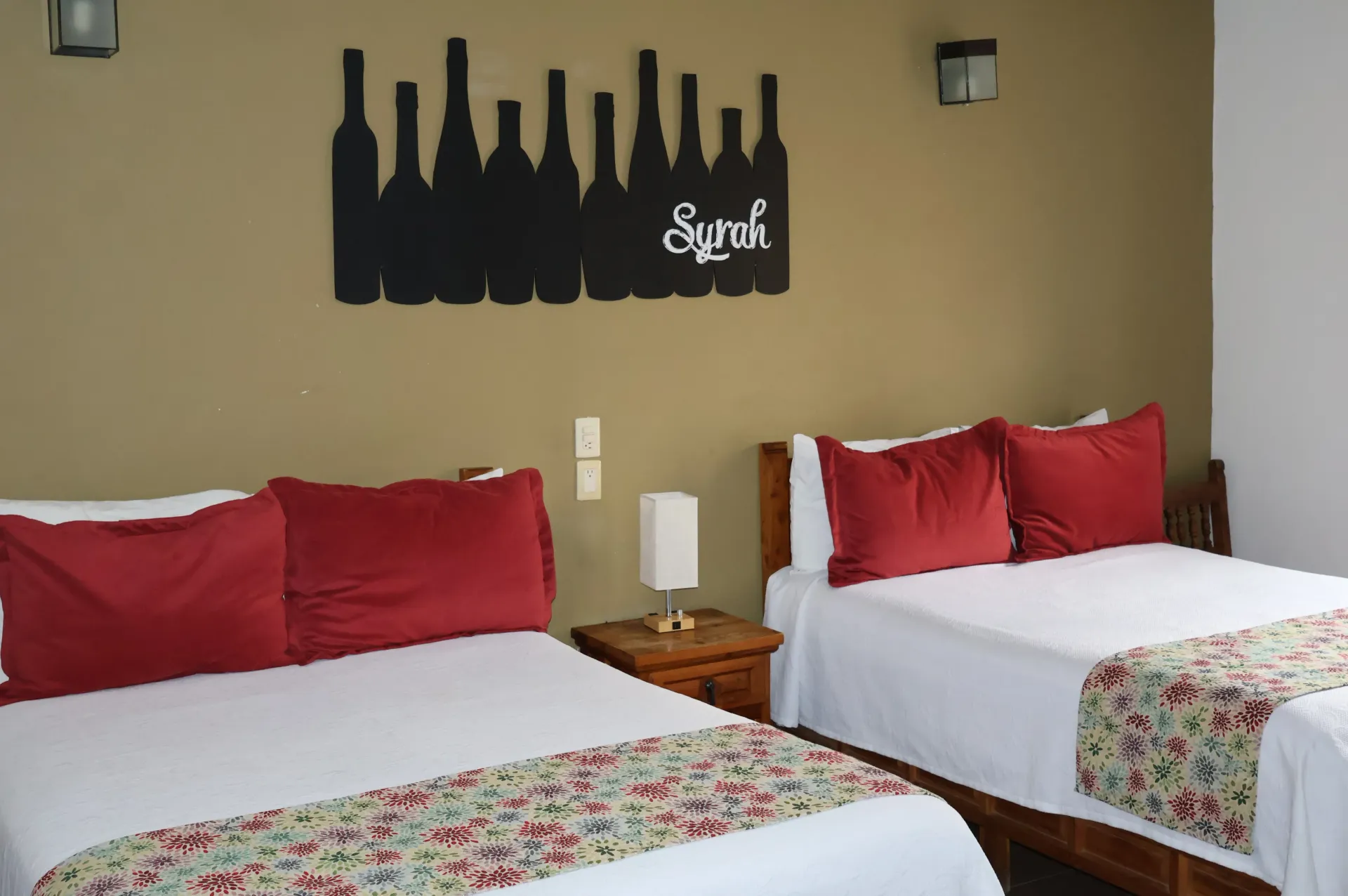 Dos camas individuales con almohadas rojas en una habitación beige con cuadros en la pared y una mesita de noche.
