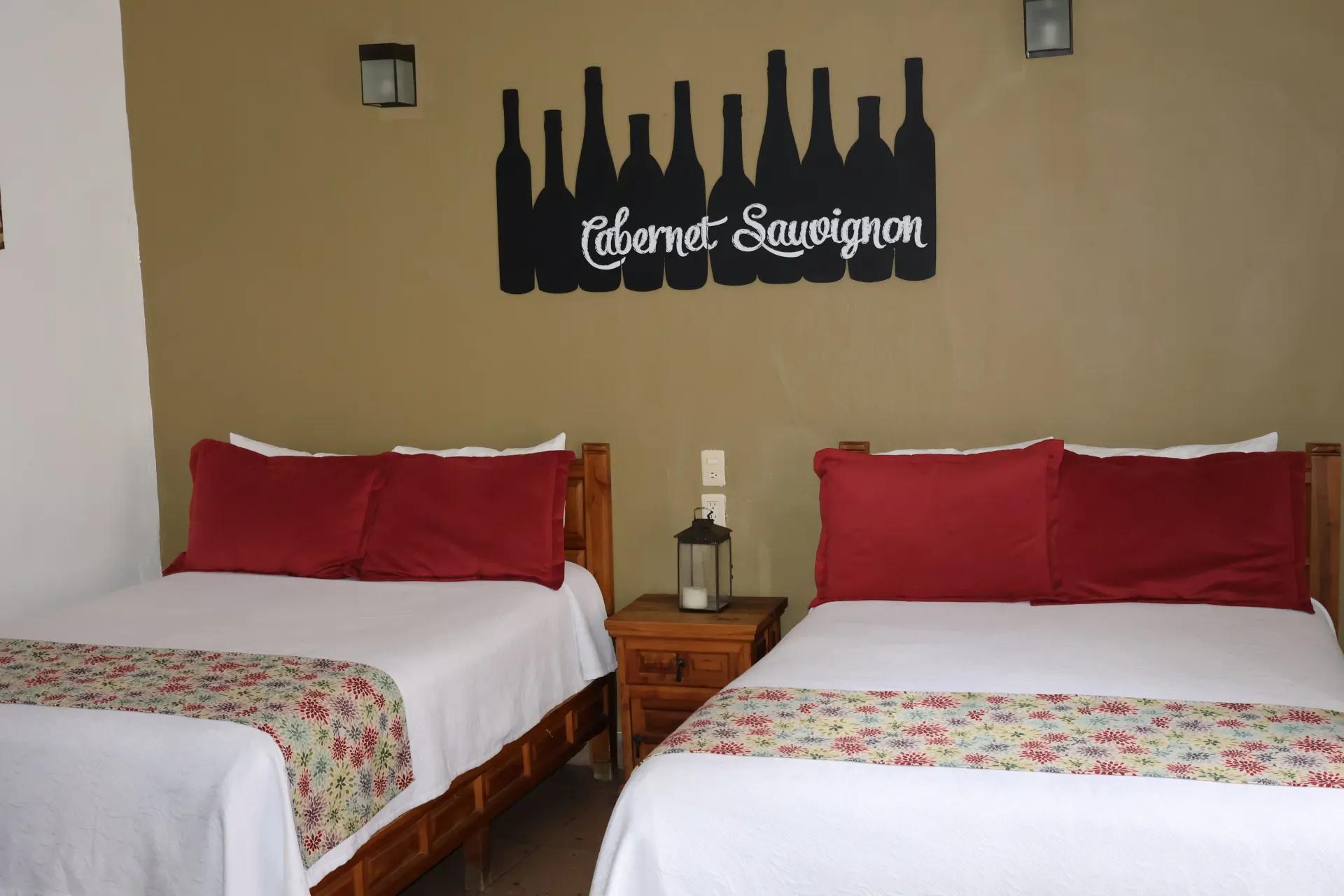 Habitación de hotel con dos camas individuales, almohadas rojas y colchas florales sobre una pared beige.
