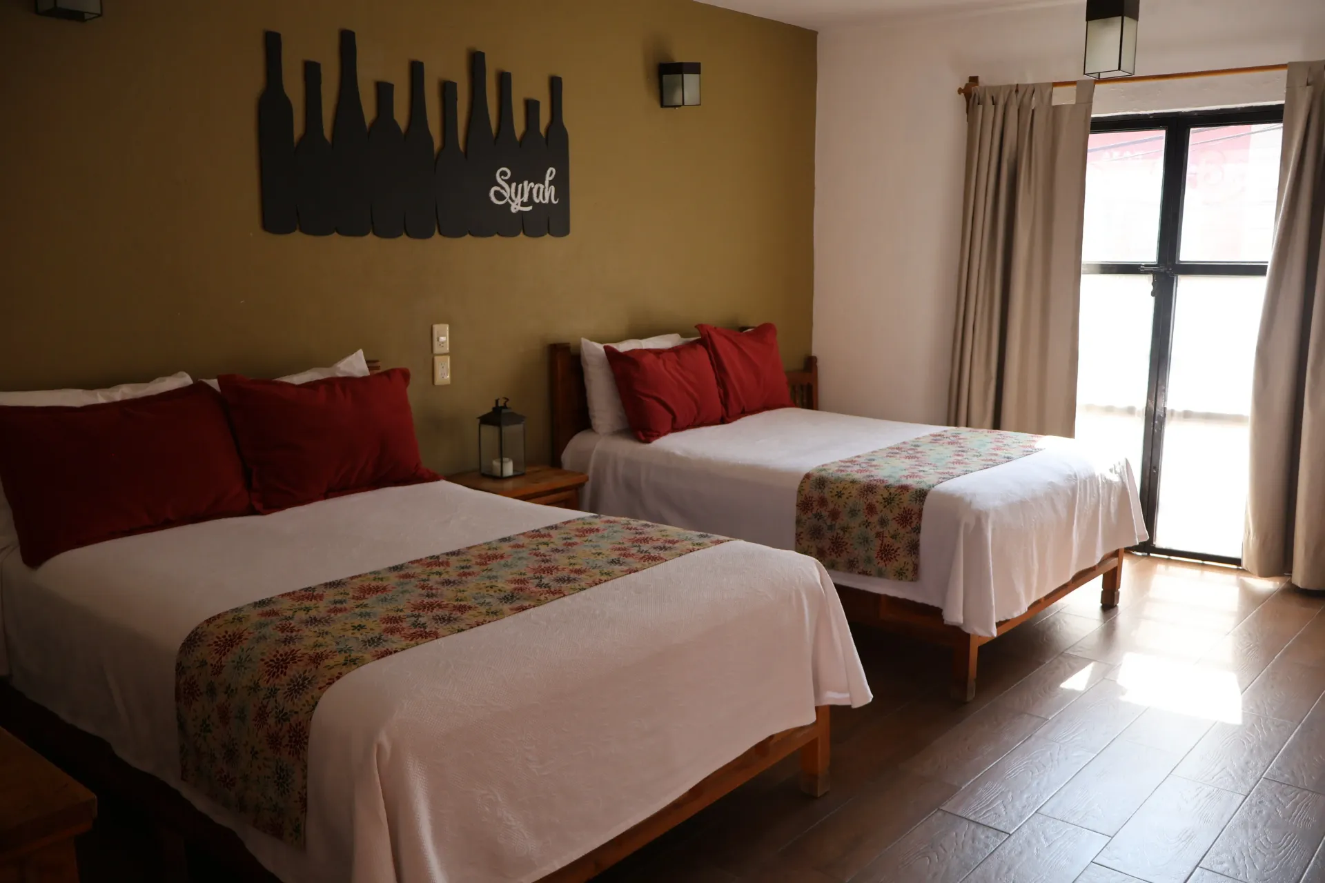 Una acogedora habitación con dos camas individuales, almohadas rojas, paredes beige y luz natural que entra por la ventana.