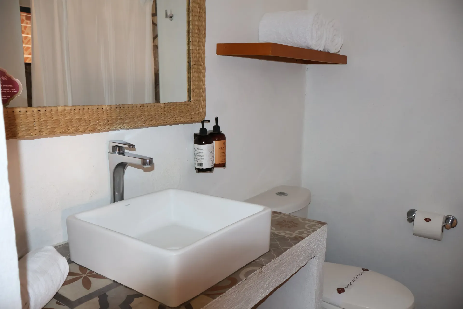 Baño moderno con lavabo cuadrado, espejo, toallas e inodoro en un interior blanco y gris.