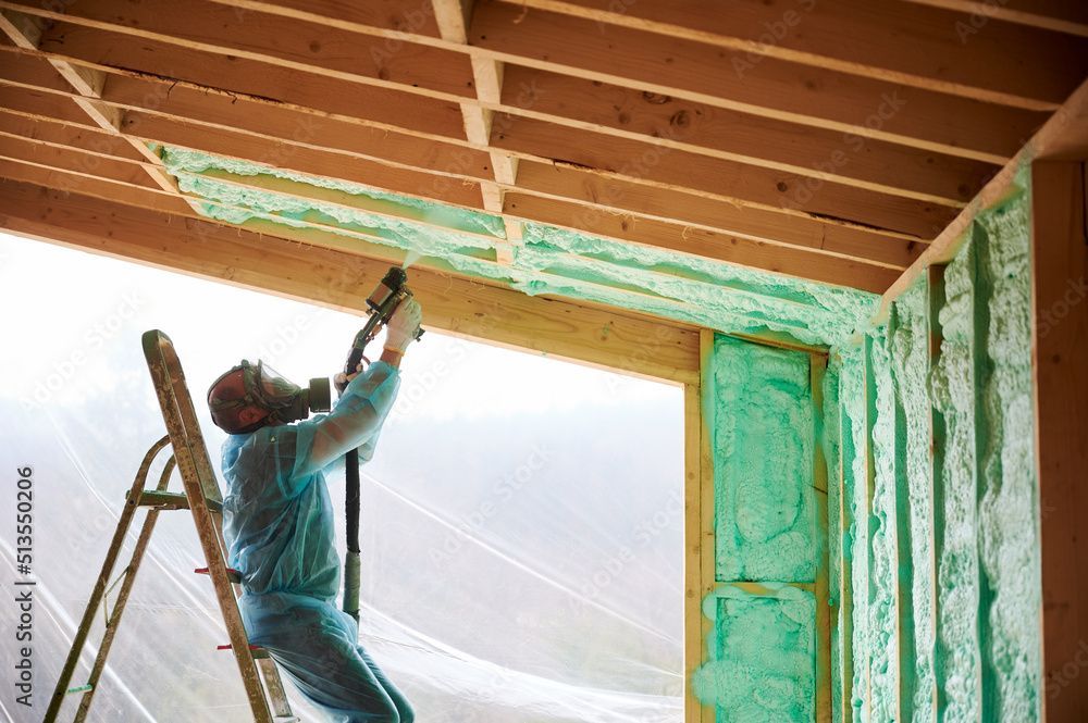 Person applying spray foam insulation to ceiling and wall of a wooden structure while wearing protective gear. Person applying spray foam insulation to ceiling and wall of a wooden structure while wearing protective gear.
