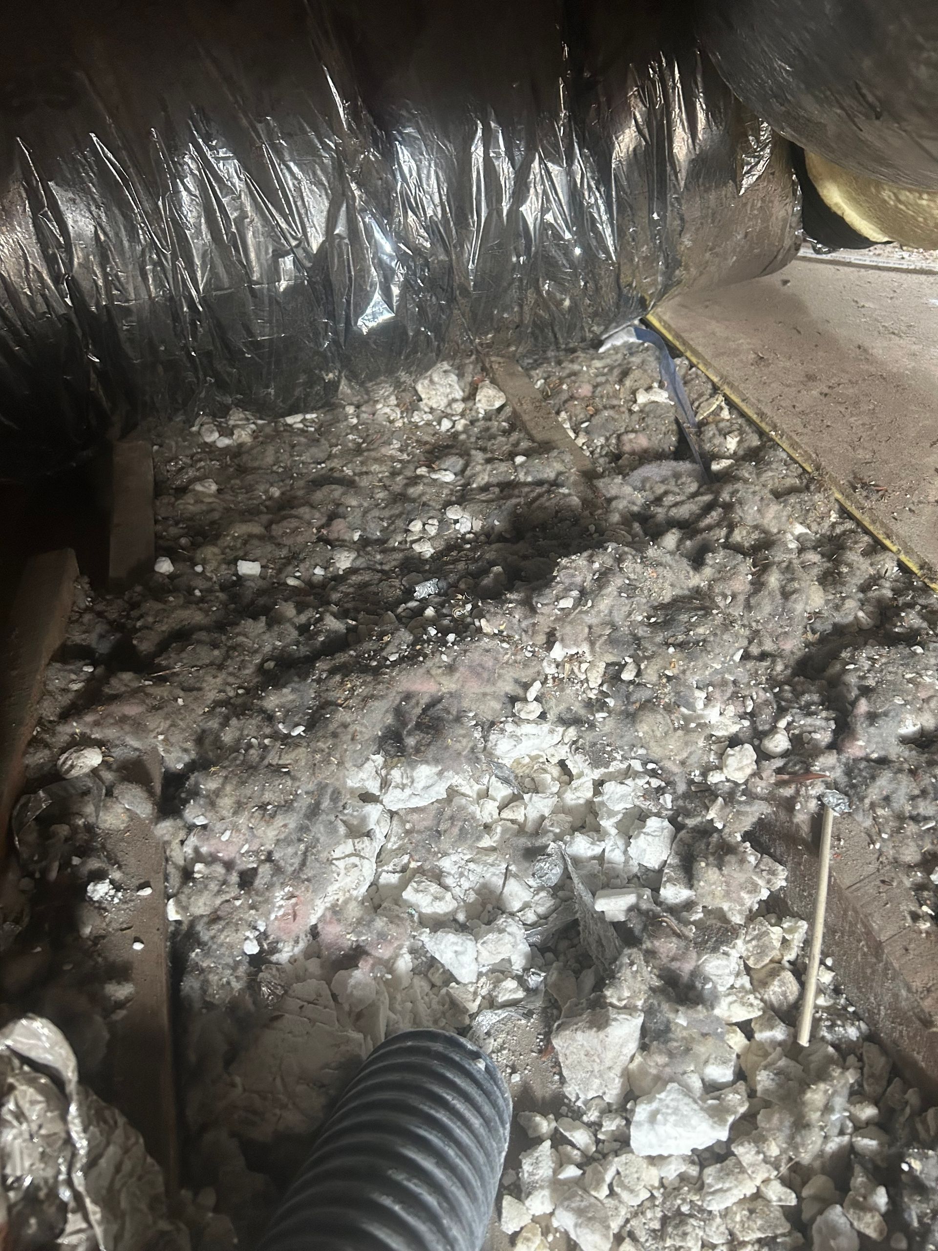 Debris and insulation inside a dark attic with a flexible duct and wood beams.
