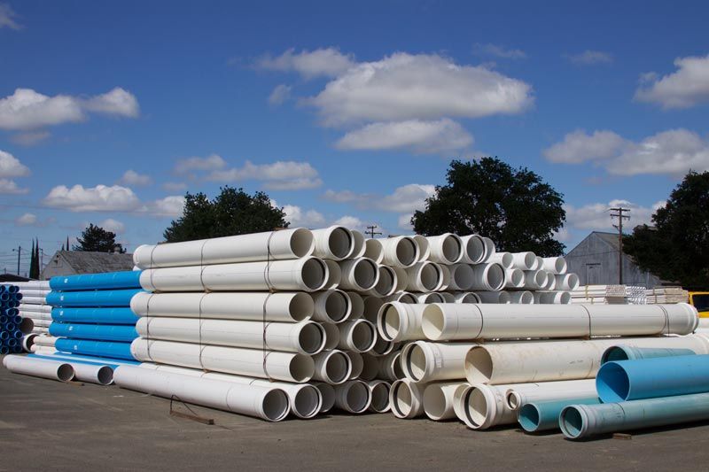 Piles of white and blue PVC pipes stacked outdoors against a cloudy blue sky.