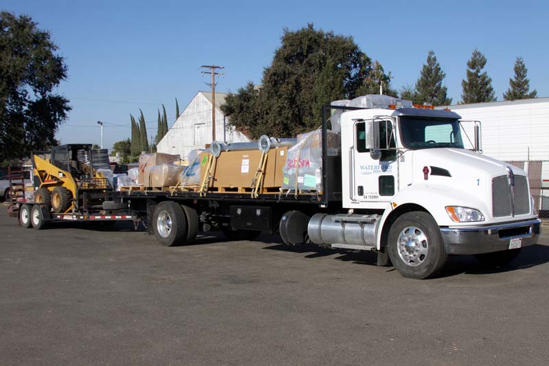White truck with flatbed carrying cargo and pulling a trailer with a yellow construction machine, parked outdoors.