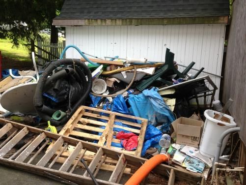 Pile of junk and debris in front of a white shed. Includes pallets, ladder, hoses, and other discarded items.