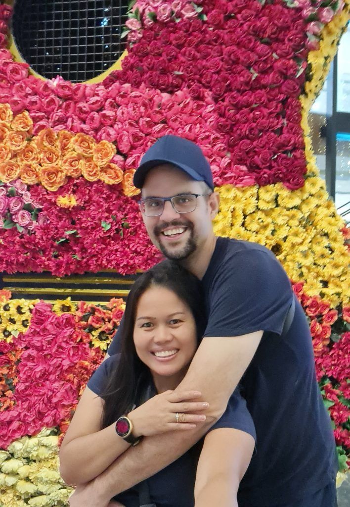 Couple smiling, hugging in front of floral guitar display; pink, orange, yellow, and red flowers.