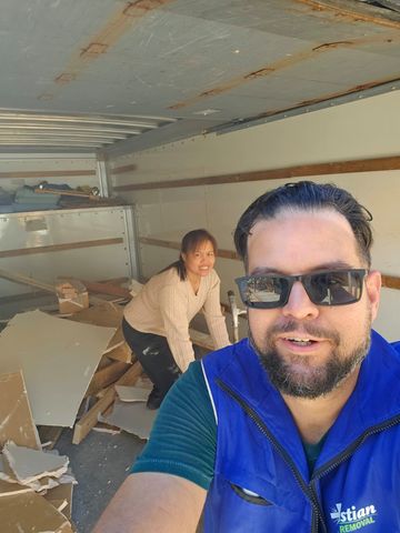A person in a blue vest takes a selfie with a smiling person crouching behind them in a trailer filled with debris.