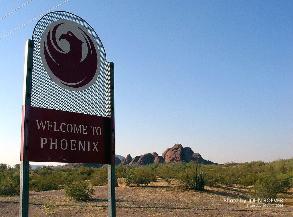Sign welcoming visitors to Phoenix with a red Phoenix bird emblem; rocky desert backdrop.