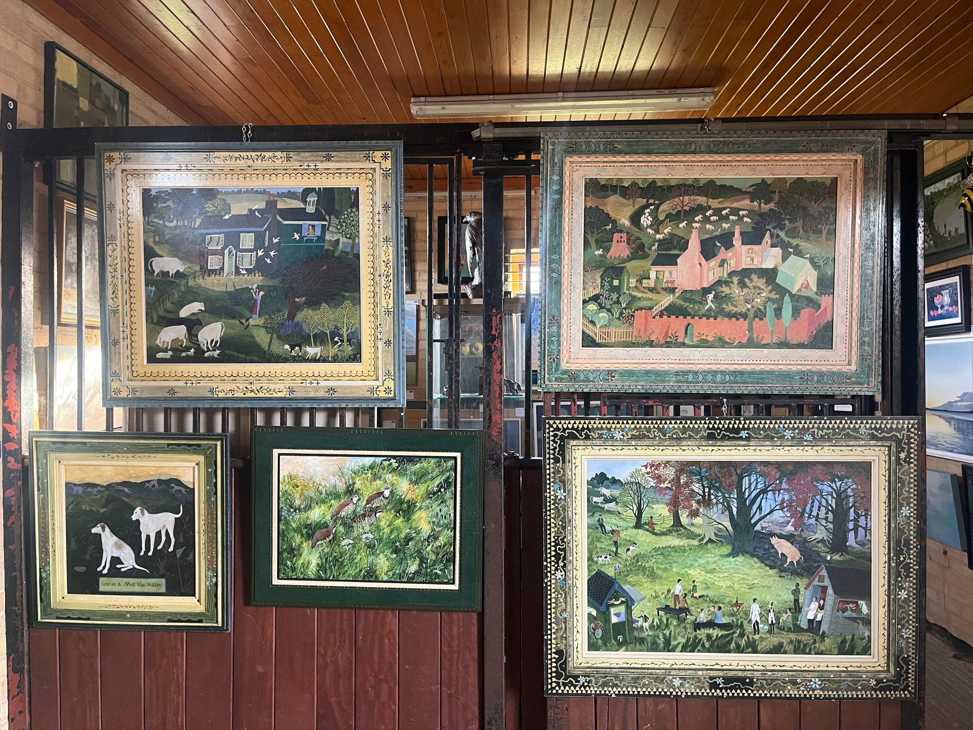 Paintings in ornate frames hang on a wooden wall. Scenes of nature, animals, and buildings.