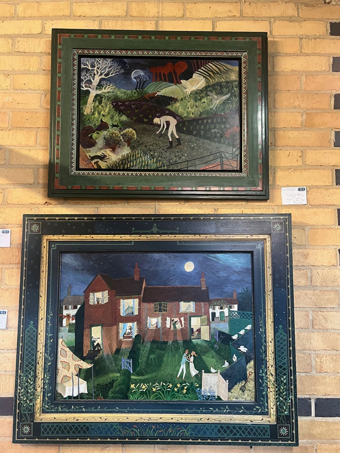 Two framed paintings on a brick wall: a night scene of a house and a landscape with a figure. Both have ornate green frames.