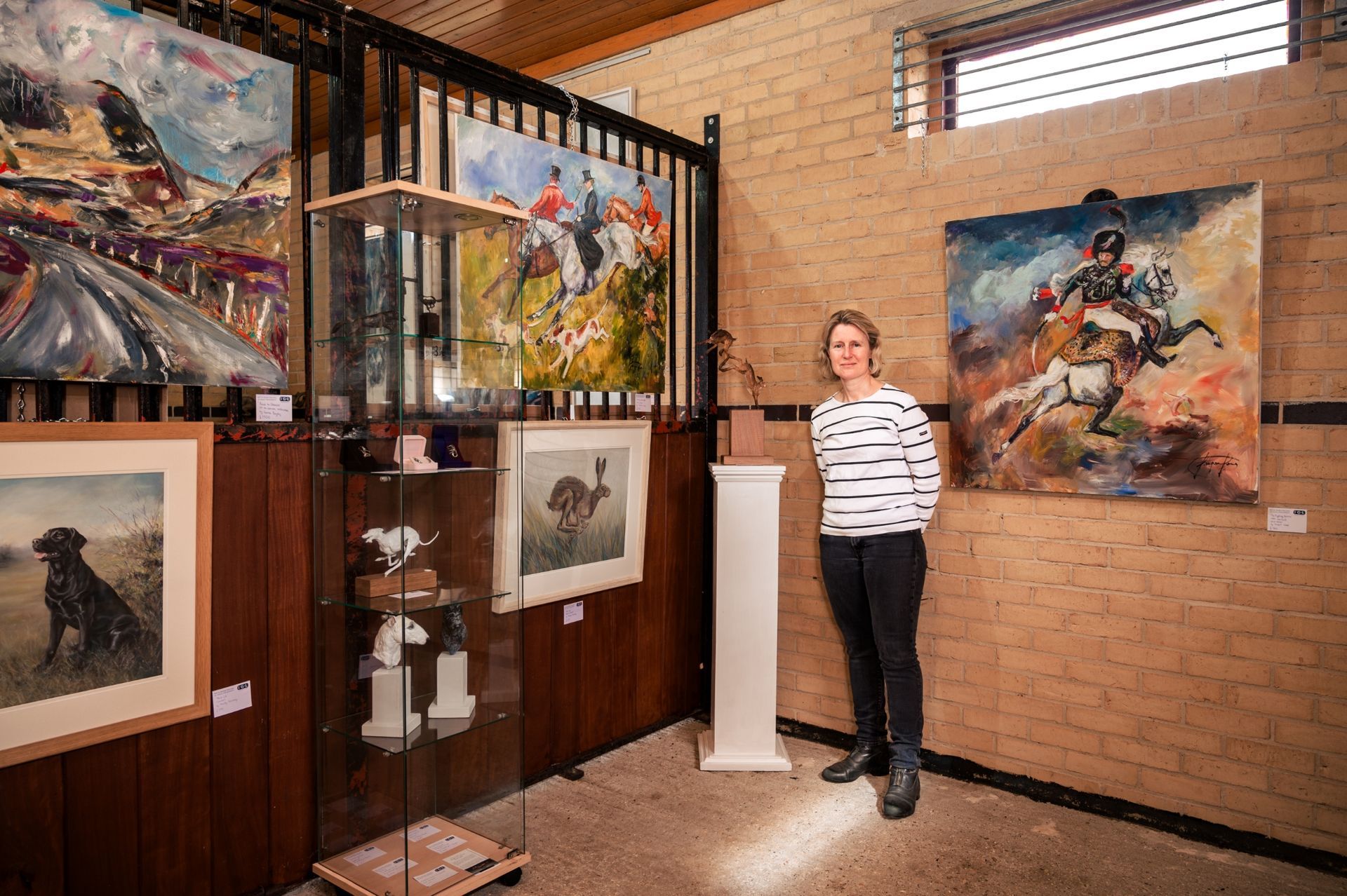 Person in gallery, surrounded by paintings of horses. Brick wall, wood and glass display.