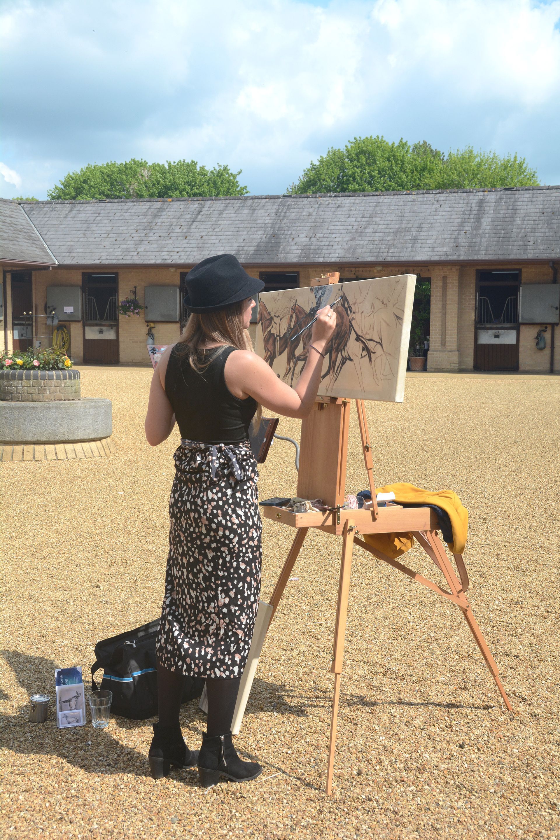 Woman in hat painting outdoors on easel; pale gravel yard with building in background.