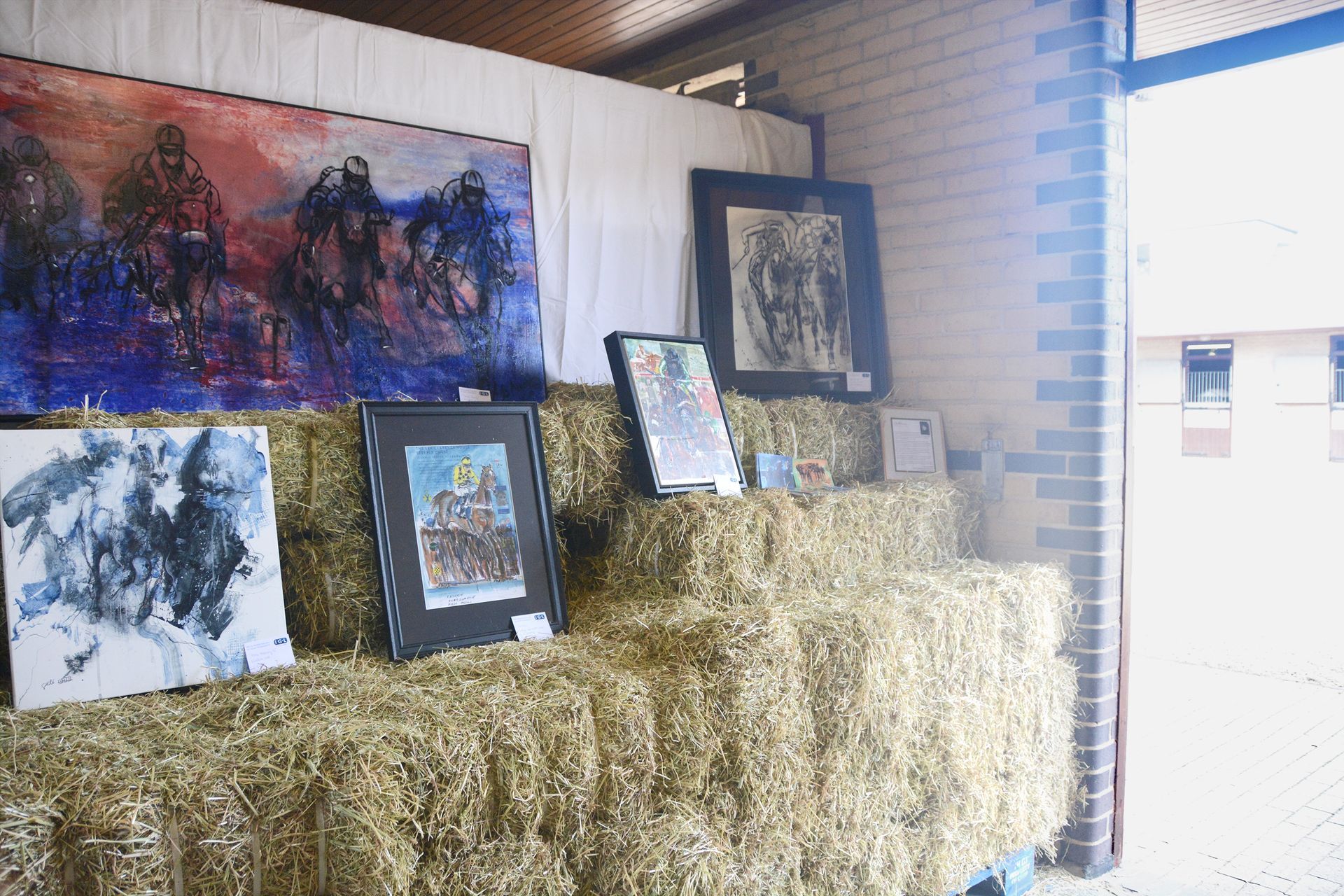 Art display on hay bales, paintings of people and horses with abstract, vibrant colors.
