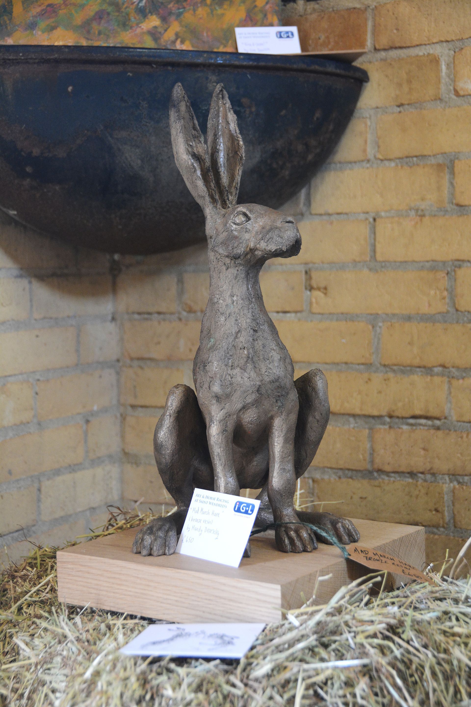 Bronze hare sculpture on wood base, seated upright, against a brick wall with a pot above and hay below.