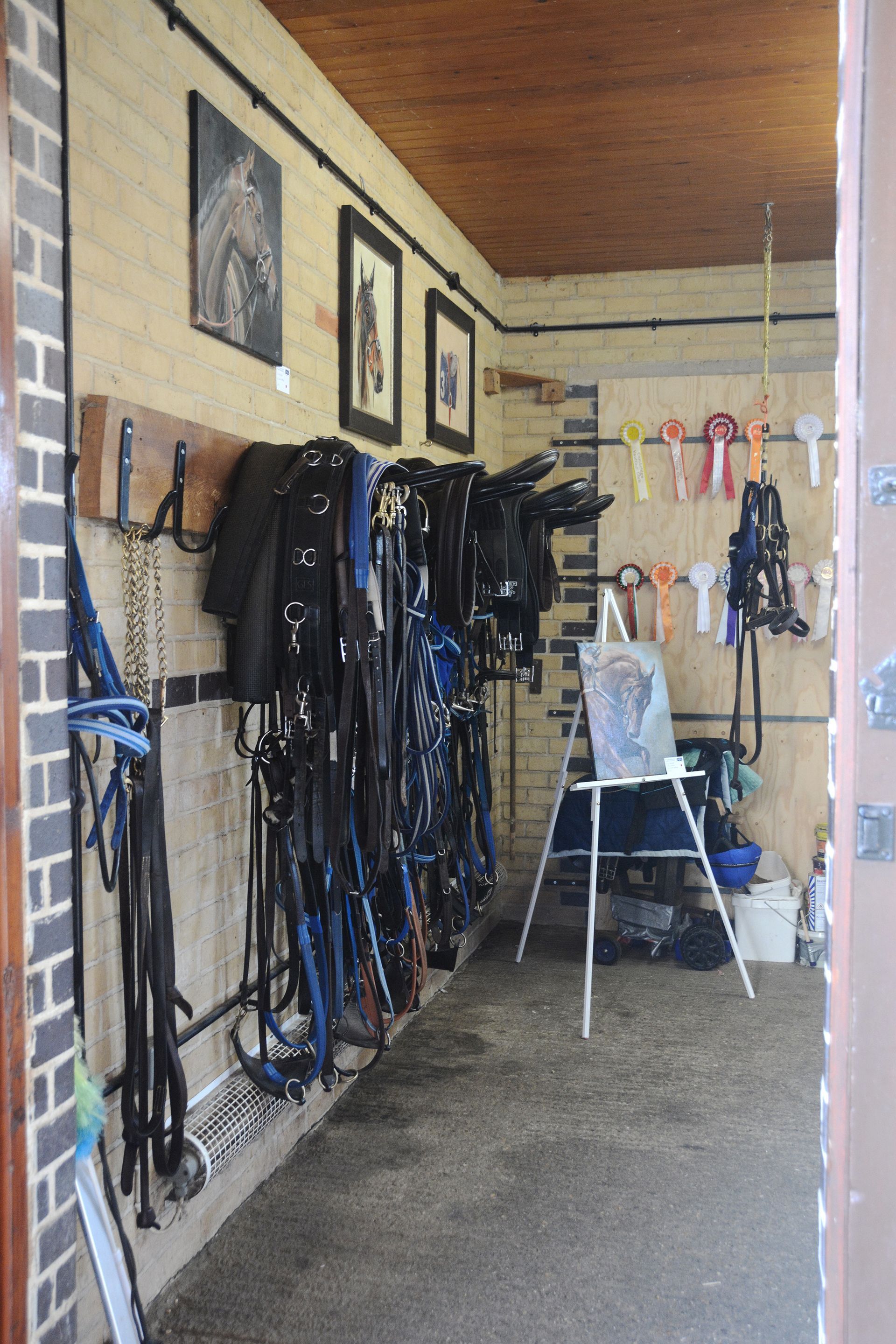 Tack room with hanging bridles, saddles, and equipment. Paintings, a painting easel, and ribbons are also visible.