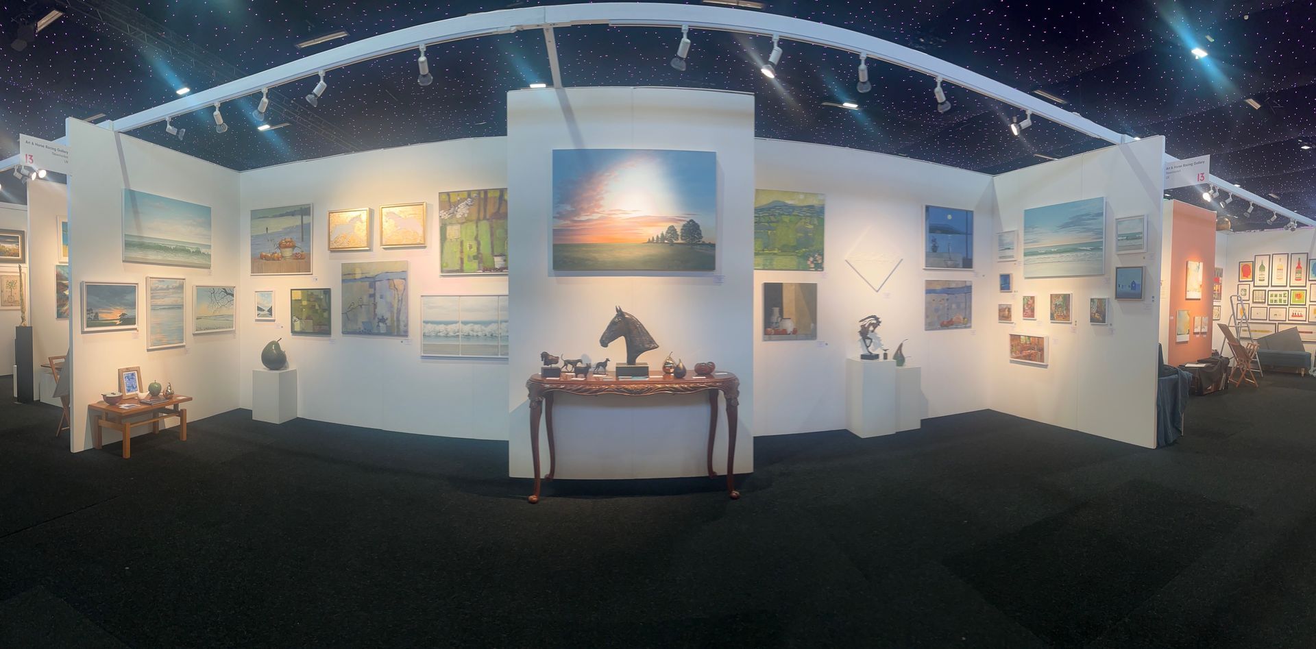 Art gallery display with framed paintings and sculptures. White walls, black carpet, overhead lighting.