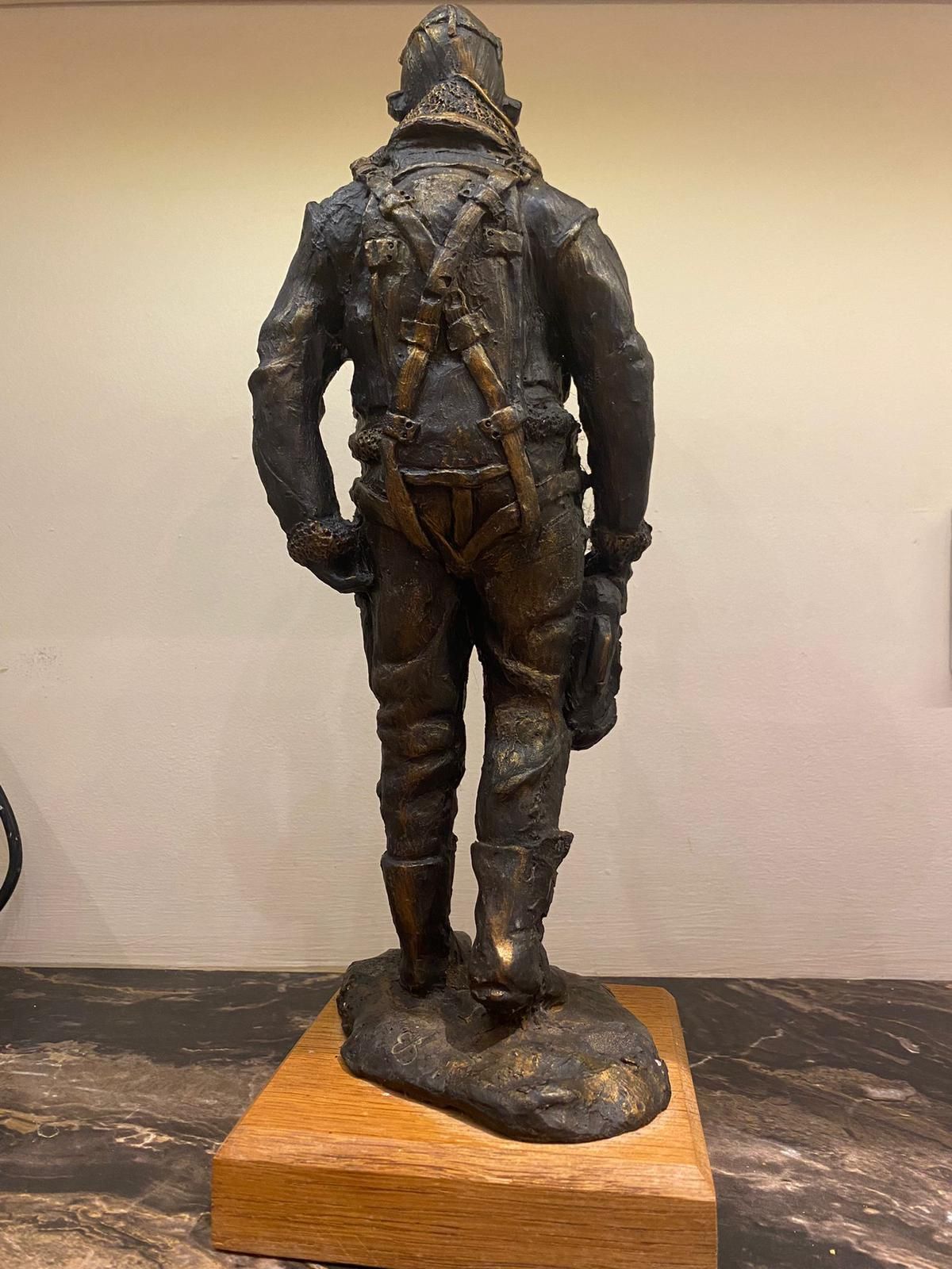 Bronze statue of a pilot from the rear, with detailed flight suit, standing on wooden base.