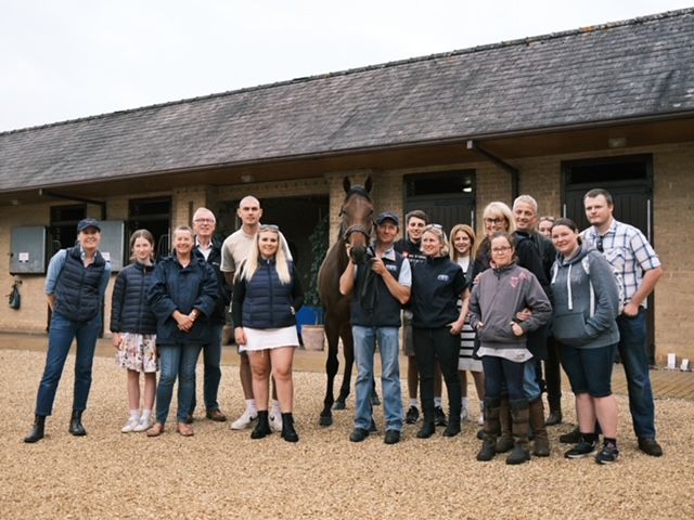 Group of people pose with a horse outside a stable. Some are smiling.