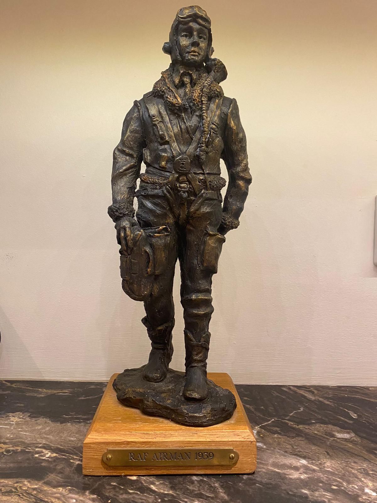Bronze statue of a pilot in a flight suit, standing on a wooden base, indoor setting.
