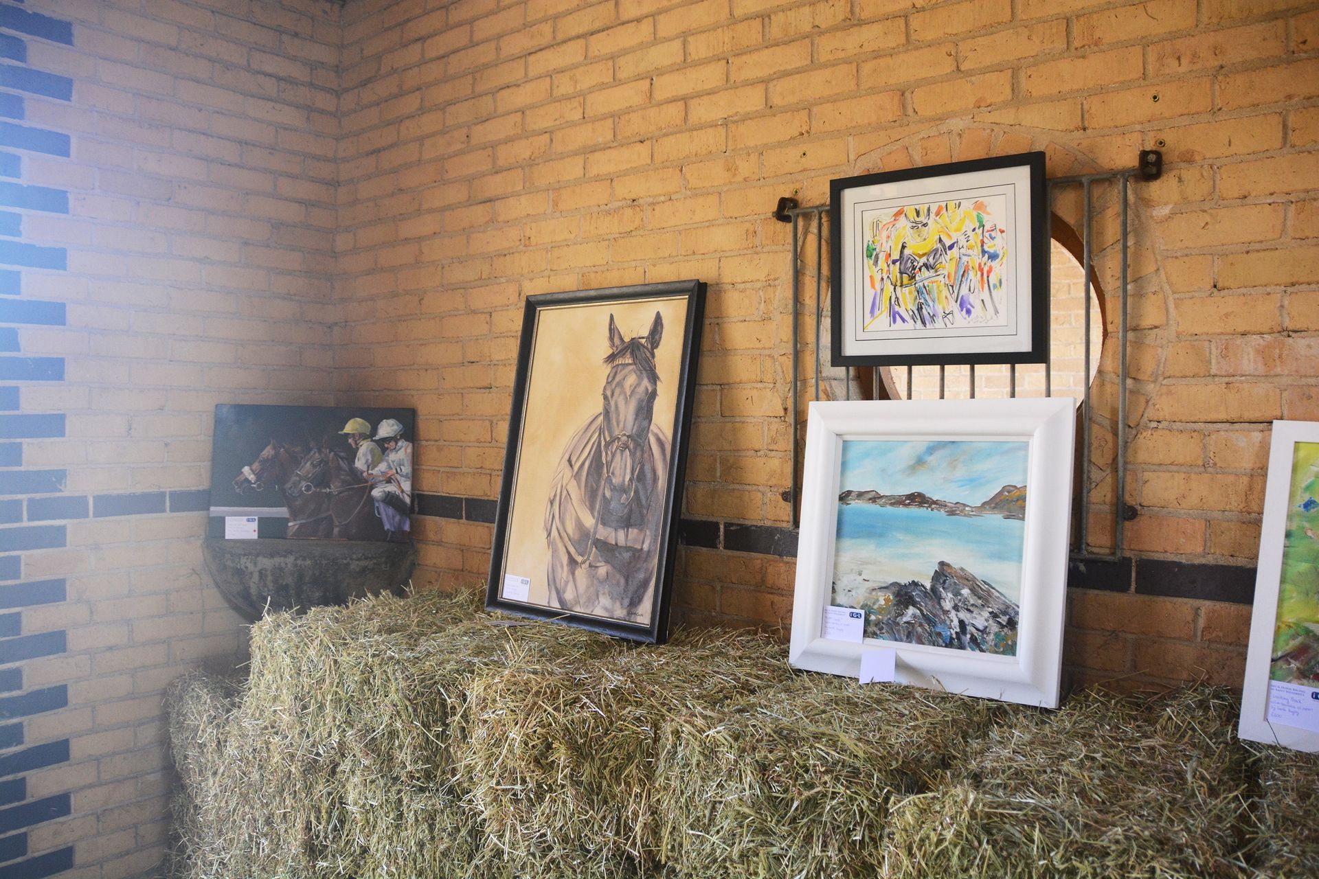 Artwork display: horse portrait, abstract art, landscape paintings on hay bales against a brick wall.