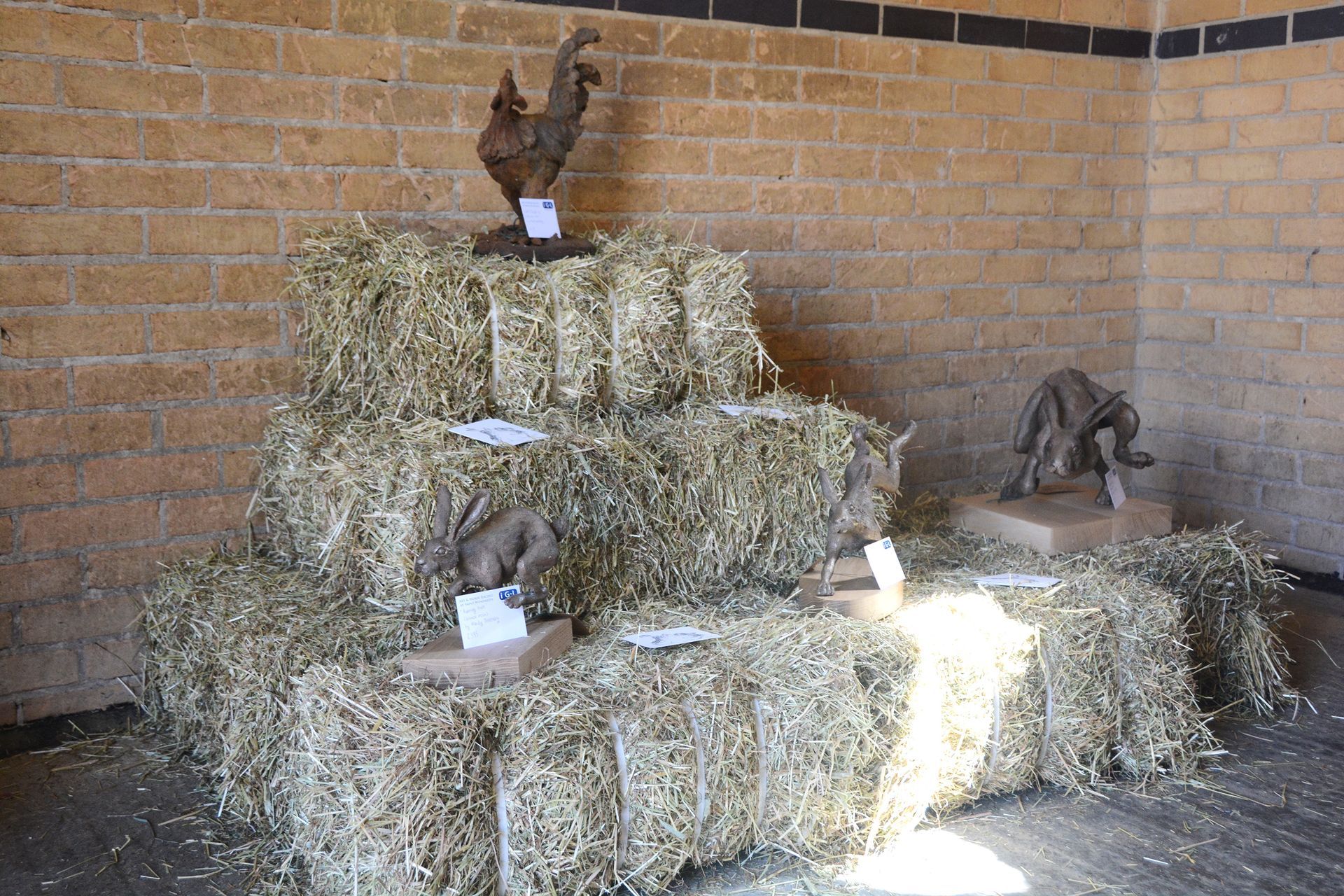 Hay bales tiered display with sculptures of a rooster and dog in a brick-walled interior.