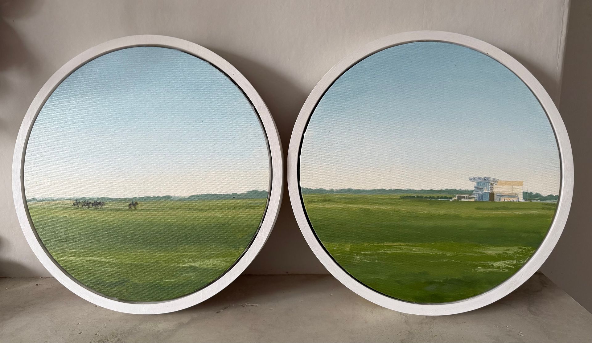 Two round paintings: green fields under a blue sky, one with a building, both in white frames.