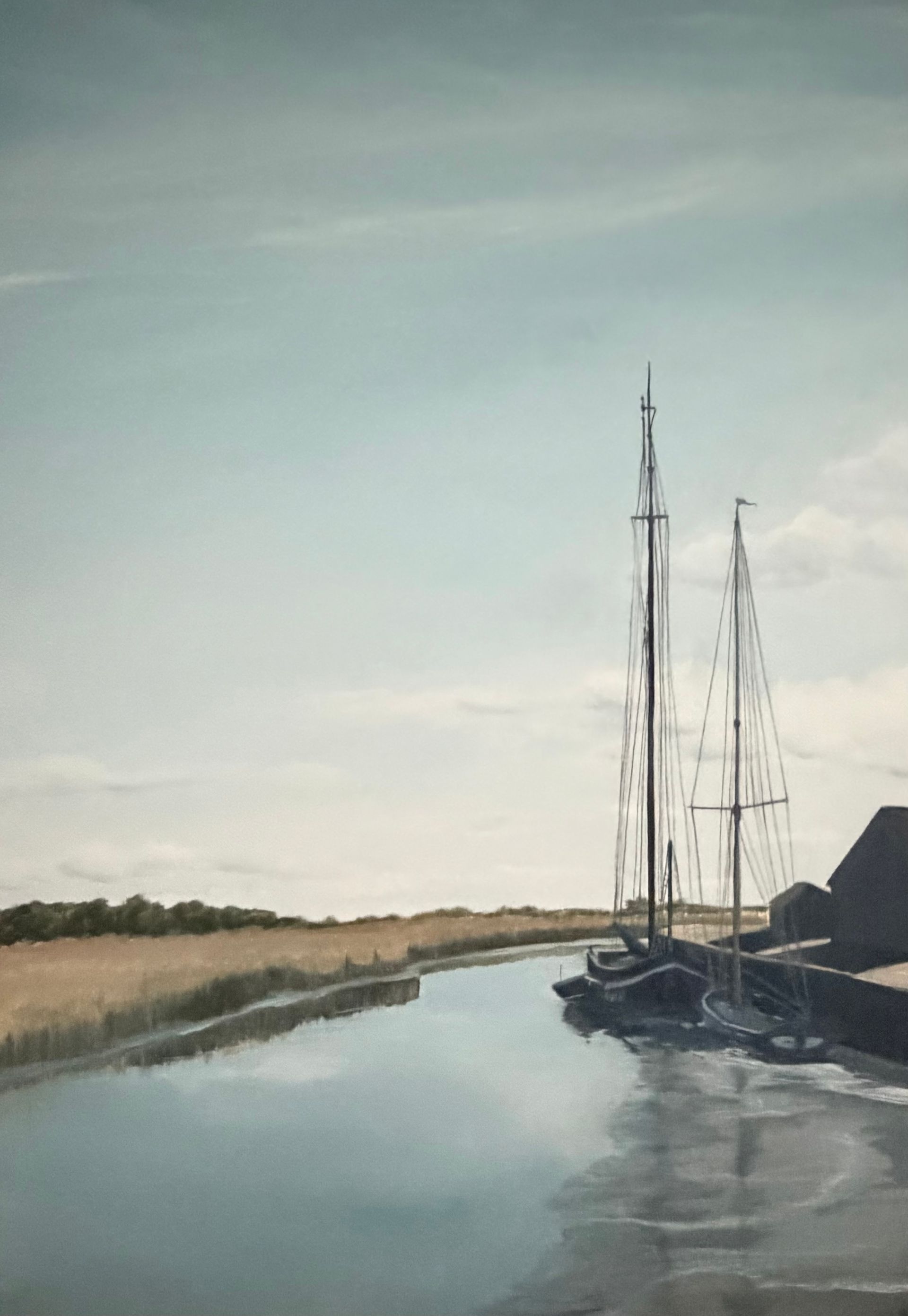 Sailing boats docked in a waterway on a sunny day. Tall masts reflect in the calm water with grassy banks.