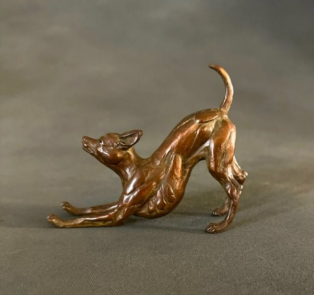 Bronze sculpture of a small dog, stretching forward with raised tail, on a gray surface.