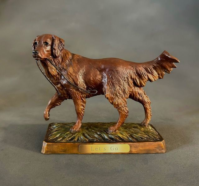 Bronze sculpture of a golden retriever holding a leash in its mouth, standing on a grassy base.