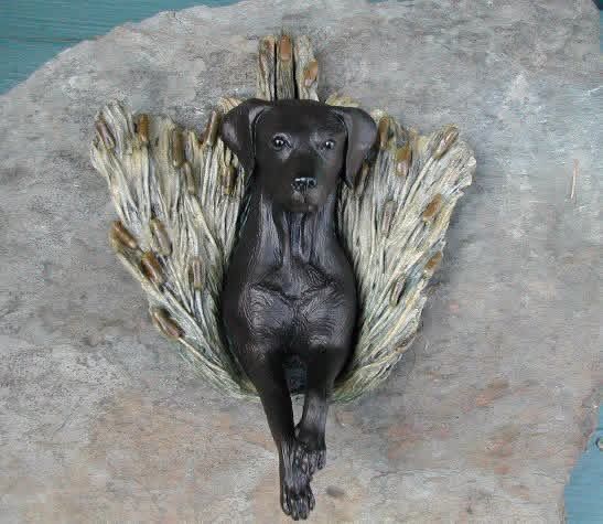 Black dog sculpture nestled in textured, light-brown material, mounted on a gray surface.