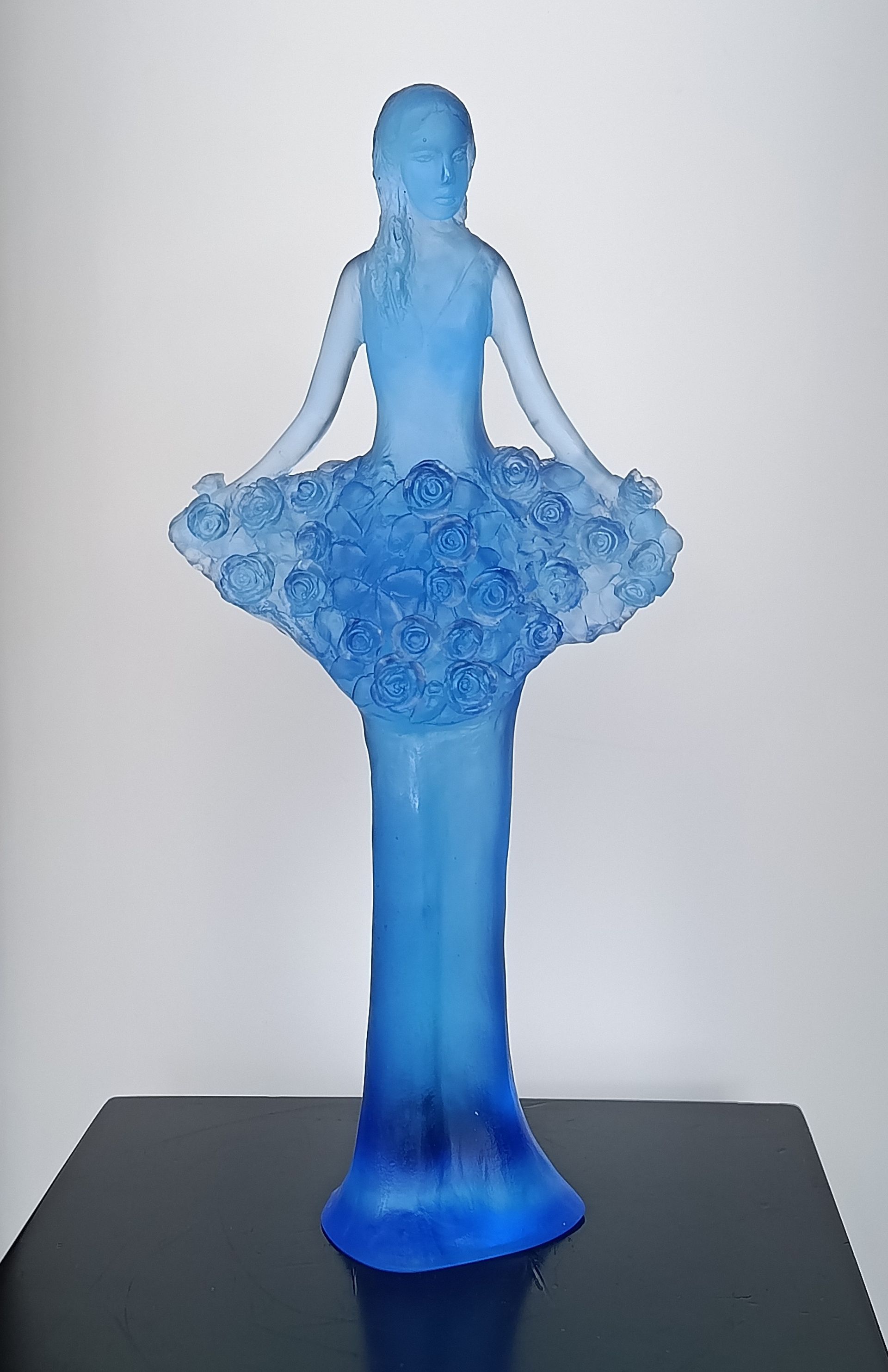 A blue glass sculpture of a figure wearing a floral-textured dress, standing on a dark base against a white background.