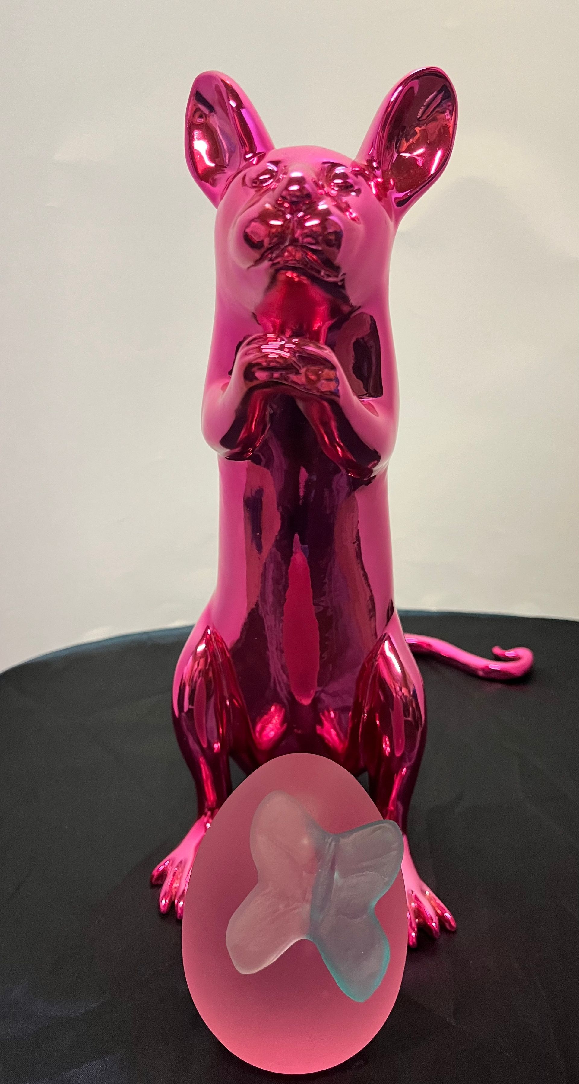 A shiny, metallic pink rat sculpture stands next to a frosted pink egg-shaped object topped with a small, pale blue flower.