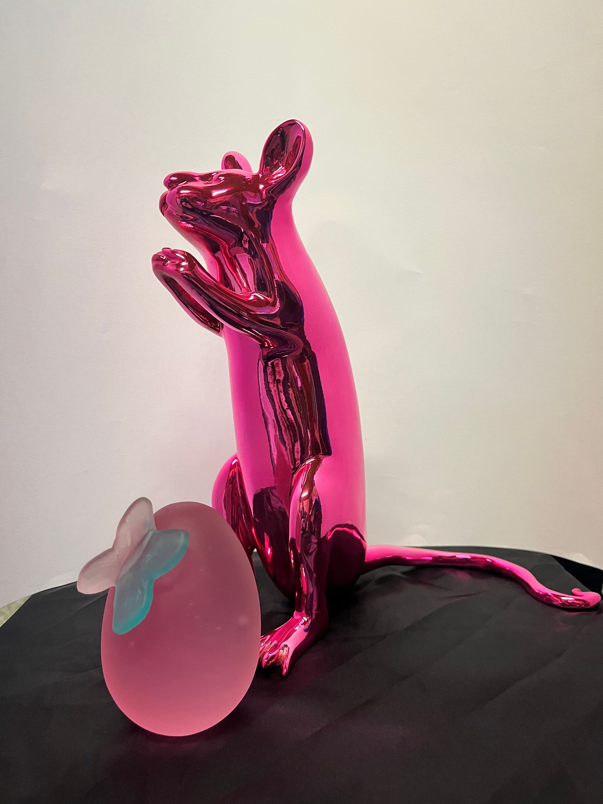 A shiny, metallic pink rat sculpture stands next to a frosted pink egg-shaped object topped with a small, pale blue flower.